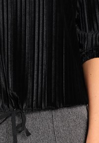 Schwarzes Samtoberteil mit vertikalen Falten, gerafften Ärmeln und einem Bindeband am Saum. Getragen über einem grauen strukturierten Stoff.