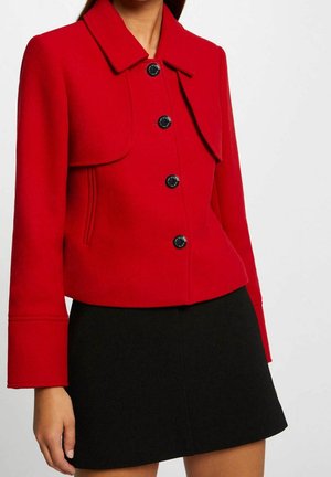 Donna che indossa una giacca aderente rossa con quattro bottoni neri e una gonna nera, mostrata dalla metà del collo fino alla metà della coscia su uno sfondo semplice.