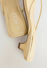 Sandali con tacco in pelle patent beige, con punta rotonda e tacco a blocco basso. Presentano un logo impresso e una soletta testurizzata per il comfort.