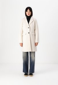Beige villasekoituksesta valmistettu takki, jossa on viiltohaarukka, kaksi etutaskua ja yksittäinen nappisulku, yhdistettynä vaaleisiin haalistuneisiin bootcut-farkkuihin.