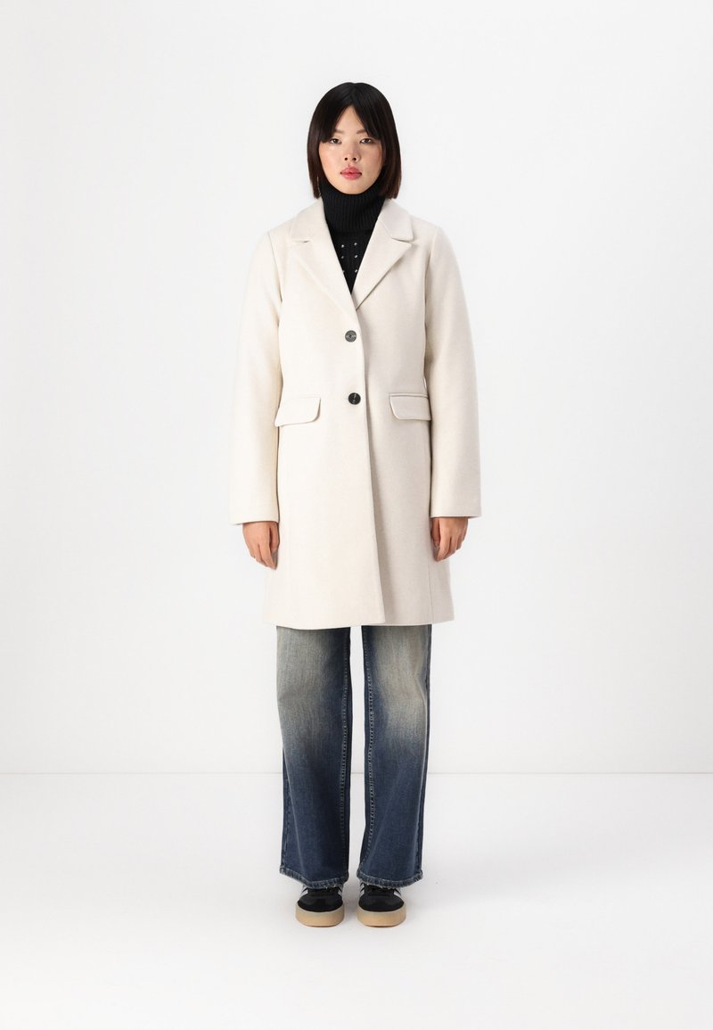 Beige villasekoituksesta valmistettu takki, jossa on viiltohaarukka, kaksi etutaskua ja yksittäinen nappisulku, yhdistettynä vaaleisiin haalistuneisiin bootcut-farkkuihin.