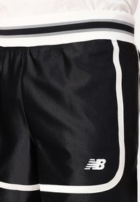 Shorts de sport noirs avec des rayures blanches sur les côtés et la taille. Logo New Balance en blanc. Matériau lisse et léger.