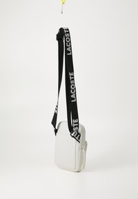 Borsa a tracolla bianca con una finitura testurizzata, dotata di una tracolla nera regolabile su cui è impresso "LACOSTE" in bianco. Chiusura con zip.