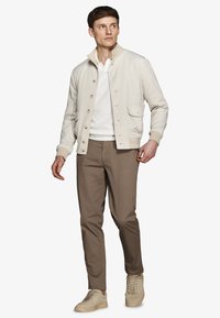 Ung mand iført beige jakke med knapper over hvid sweater, brune bukser og beige sneakers, stående med et neutralt udtryk.