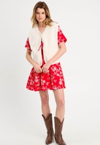 Gilet crémeux et duveteux avec un devant à boutons sur une robe fleurie rouge. Des bottes de cowboy marron complètent la tenue.