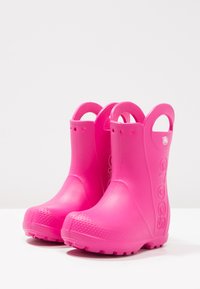 Bottes de pluie en caoutchouc rose avec une finition lisse, un bout arrondi et une semelle texturée. Les caractéristiques comprennent des poignées sur les côtés et un logo sur le côté extérieur.