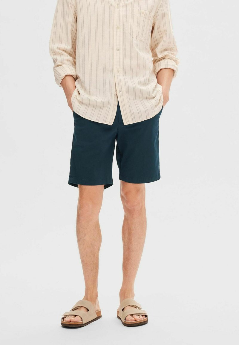 Camicia button-up a righe beige chiaro, pantaloni corti teal scuro e sandali beige con cinturini larghi. Completo estivo casual con vestibilità rilassata.