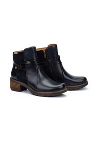 Botas de tobillo de cuero negro con punta redondeada, cremallera lateral y correa decorativa envuelta. Presentan una suela texturizada marrón y un tacón de altura media.