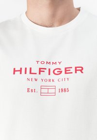 Weißes Baumwoll-T-Shirt mit bold rot gedrucktem Text: "TOMMY HILFIGER NEW YORK CITY" und "Est. 1985" sowie klassischem Rundhalsausschnitt.
