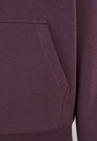 Gros plan d'une poche de sweat à capuche en tissu bordeaux avec des coutures visibles et un ourlet côtelé en bas.