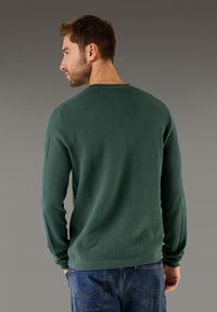 Zelená, texturovaná košile s dlouhým rukávem a kulatým výstřihem, doplněná modrými džíny. Pohled zezadu ukazuje rukávy těsně přiléhající na zápěstích.