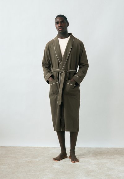 Άνδρας που στέκεται ξυπόλητος σε χαλί, φορώντας χιτώνα ελιάς με λευκή επένδυση, δεμένο στη μέση πάνω από λευκό πουκάμισο, σε απλό φόντο.