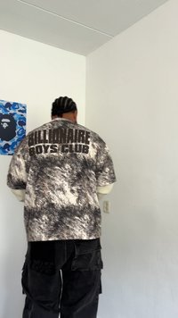 Person with braided hair wearing a black and white Billionaire Boys Club shirt, standing against a white wall with a blue artwork nearby.