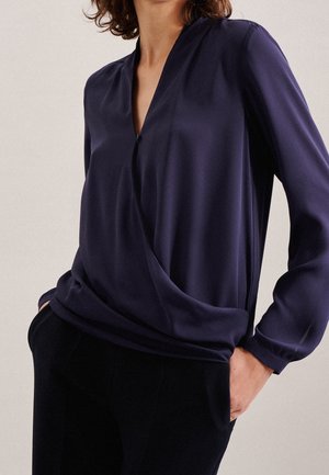 Chemisier bleu marine en tissu lisse, avec un décolleté en V, des manches longues et un design drapé, rentré dans un pantalon noir taille haute.