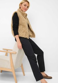 Gilet en fausse fourrure beige avec un col montant, associé à un haut noir et un pantalon noir slim. La personne est assise sur un fauteuil en velours côtelé clair.