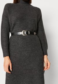 Donna che indossa un vestito a costine grigio scuro con maniche lunghe, cinstrato in vita da una cintura in pelle nera con una fibbia decorativa argentata.