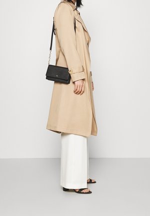 Trench coat beige avec col et boutons décoratifs, associée à un pantalon blanc à jambes larges et des sandales noires. Un petit sac noir porté en bandoulière.