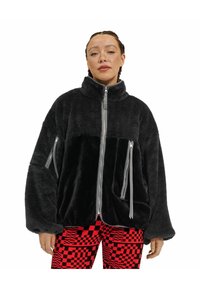 Giacca nera in pelliccia sintetica con collo alto, zip frontale completa e due tasche laterali, con dettagli in zip grigia e vestibilità comoda.