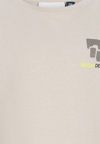 T-shirt beige con scollatura rotonda che presenta una grafica testuale nera e gialla "RETOUR" e un design a strisce nere sulla parte superiore sinistra del petto.