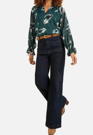 Femme portant un chemisier vert à manches longues à motifs, un jean foncé à jambes larges avec poches avant, une ceinture marron et des chaussures à talons beiges, debout les jambes croisées.