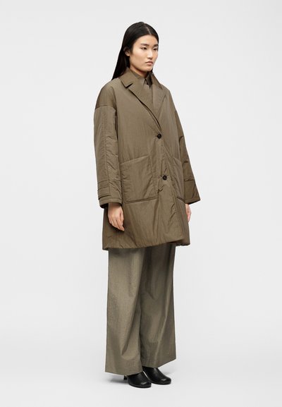 Nő áll, túlméretezett olívazöld puffadt kabátot, hozzáillő bő szárú nadrágot és fekete bokacsizmát visel, egyszerű fehér háttér előtt.