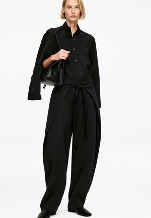 Czarna koszula na guziki z wysokim kołnierzem, zestawiona z szerokimi, luźnymi czarnymi spodniami zawiązanymi w talii. Modelka trzyma czarną torbę, ma na sobie czarne buty.