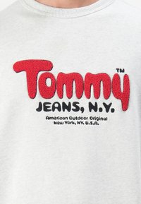 Light gray sweatshirt featuring bold, red plush embroidery of "Tommy" and black text "JEANS, N.Y." with "American Outdoor Original" below.
