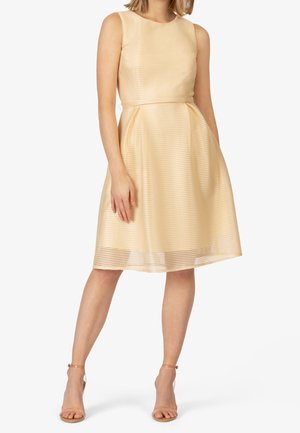 Femme portant une robe sans manches texturée de couleur or clair jusqu'aux genoux avec une jupe plissée et des escarpins beiges à bride cheville, debout devant un fond blanc.