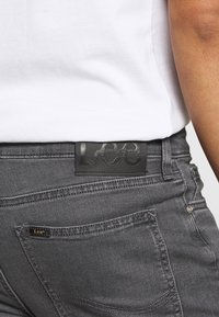 Los jeans de denim gris cuentan con un parche de cuero negro con la marca "Lee" en la cintura y costuras prominentes en los bolsillos traseros.