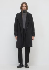 Donna con capelli lunghi che indossa un soprabito nero, un maglione a collo alto grigio a costine, pantaloni neri e mocassini neri, in piedi davanti a uno sfondo semplice.