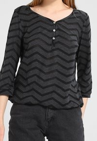 Grå långärmad topp med ett svart chevronmönster, med en V-ringning och tre knappar framtill. Mjuk tyg med avslappnad passform.