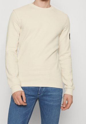 Mann trägt cremefarbenes, strukturiertes Langarmshirt mit Rundhalsausschnitt und blaue Jeans, steht vor einem schlichten hellen Hintergrund.