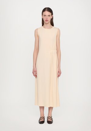 Robe beige sans manches avec détail froncé sur le côté, à hauteur des genoux, fabriquée en tissu doux. Associée à des chaussures plates foncées.