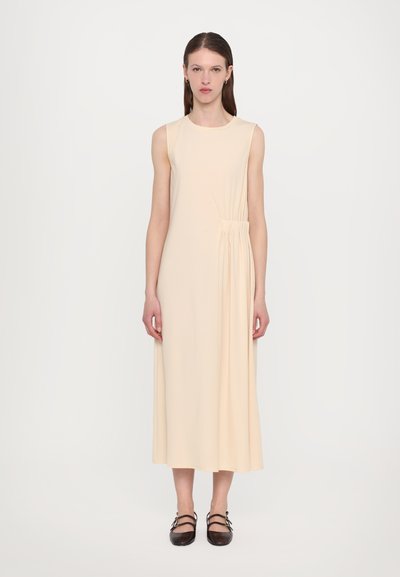 Abito beige senza maniche con dettaglio arricciato sul lato, lungo fino al ginocchio, realizzato in morbido tessuto. Abbinato a scarpe flat scure.