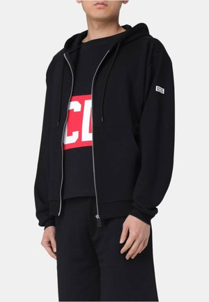 Persona che indossa una felpa nera con zip e il logo GCDS sulla manica, con una maglietta nera sotto con testo audace rosso e bianco.