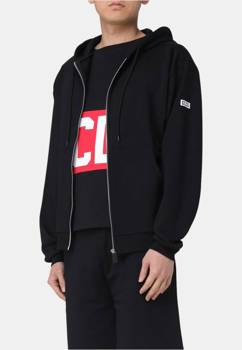 Osoba ubrana w czarną bluzę z kapturem zapinaną na suwak z logo GCDS na rękawie oraz czarną koszulkę z czerwono-białym, wyraźnym napisem pod spodem.