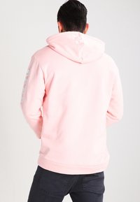 Sudadera rosa con capucha ajustable y puños acanalados. Presenta un logo texturizado en la parte trasera y detalles impresos a lo largo de la manga izquierda.