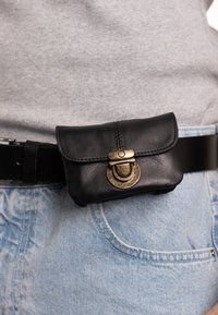 Black leather pouch with curved flap closure and brass hardware, attached to a dark belt, worn over light denim jeans.