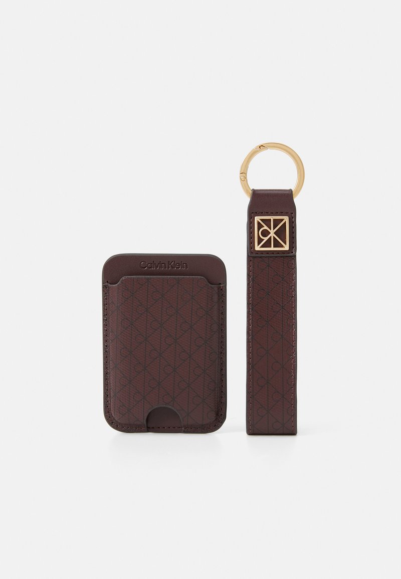 Porta-cartões de couro castanho com um padrão geométrico em relevo, acompanhado por uma alça com um anel dourado e um detalhe decorativo com logotipo.
