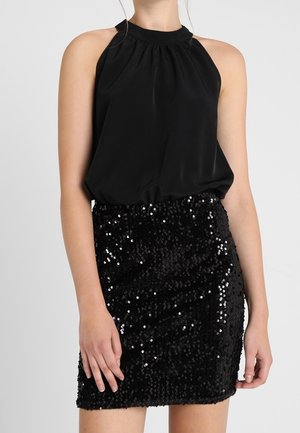 Femme portant un top noir sans manches à col licou et une mini-jupe noire à paillettes sur un fond uni.