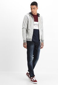 Grå hettejakke med glidelås, lagd over en burgunder og marineblå logo t-skjorte, kombinert med mørke jeans og røde sneakers.