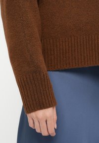Detailní záběr ruky v hnědé žebrované svetré, která odpočívá na modré sukni nebo kalhotách.