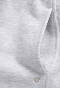 Grauer Sweatshirtstoff mit einer weichen Textur, ausgestattet mit einer vorderen Tasche und einem kleinen silbernen Logoakzent am unteren Rand.