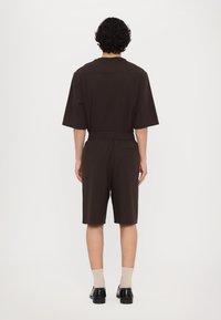 Brown short-sleeve jumpsuit with a relaxed fit, featuring an elastic waistband and side pockets. Plain texture, no visible patterns or embellishments.