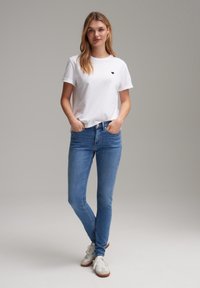 Witte katoenen t-shirt met een zwart hartlogo, gecombineerd met lichtblauwe slim-fit jeans en witte sneakers met bruine accenten.