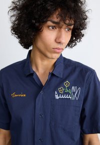 Marineblauwe kortebloes met een borstzak, versierd met kleurrijke geborduurde bloemen, een vork, een mes en een potlood, met de tekst "Service".