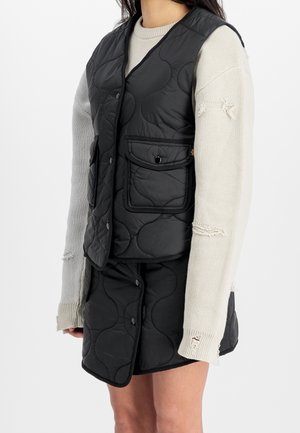 Frau trägt eine schwarze gesteppte Weste und einen Rock mit einem beigefarbenen, ausgewaschenen Strickpullover vor einem schlichten weißen Hintergrund.