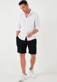 Witte langarm overhemdbloes, zwarte gestructureerde shorts, witte sneakers, staande pose, handen in de zakken, neutrale achtergrond.