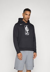 Čierna mikina Nike s predným vreckom, obsahuje logo NBA, kapucňu so šnúrkami, rebrované manžety a spodok, nosená so svetlošedými šortkami.