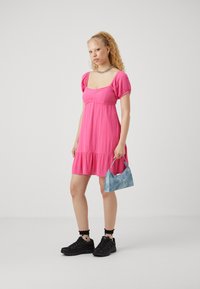 Rožinė su pūstomis rankovėmis suknelė su prigludusia liemenėle ir sluoksniuota sijonu. Su ja derinama maža mėlyna rankinė ir juodi sportbačiai su tekstūruotais padu.